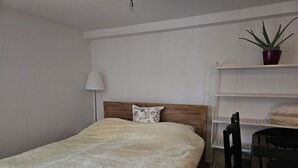 A bedroom with a bed, a wooden headboard, a bedside lamp, a potted plant, and a shelf with towels.