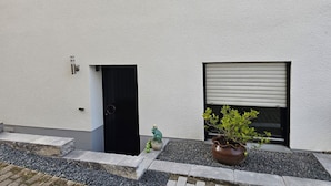 A building facade with a textured white wall, a black door, a window with a roller blind, and a potted plant with a frog statue.