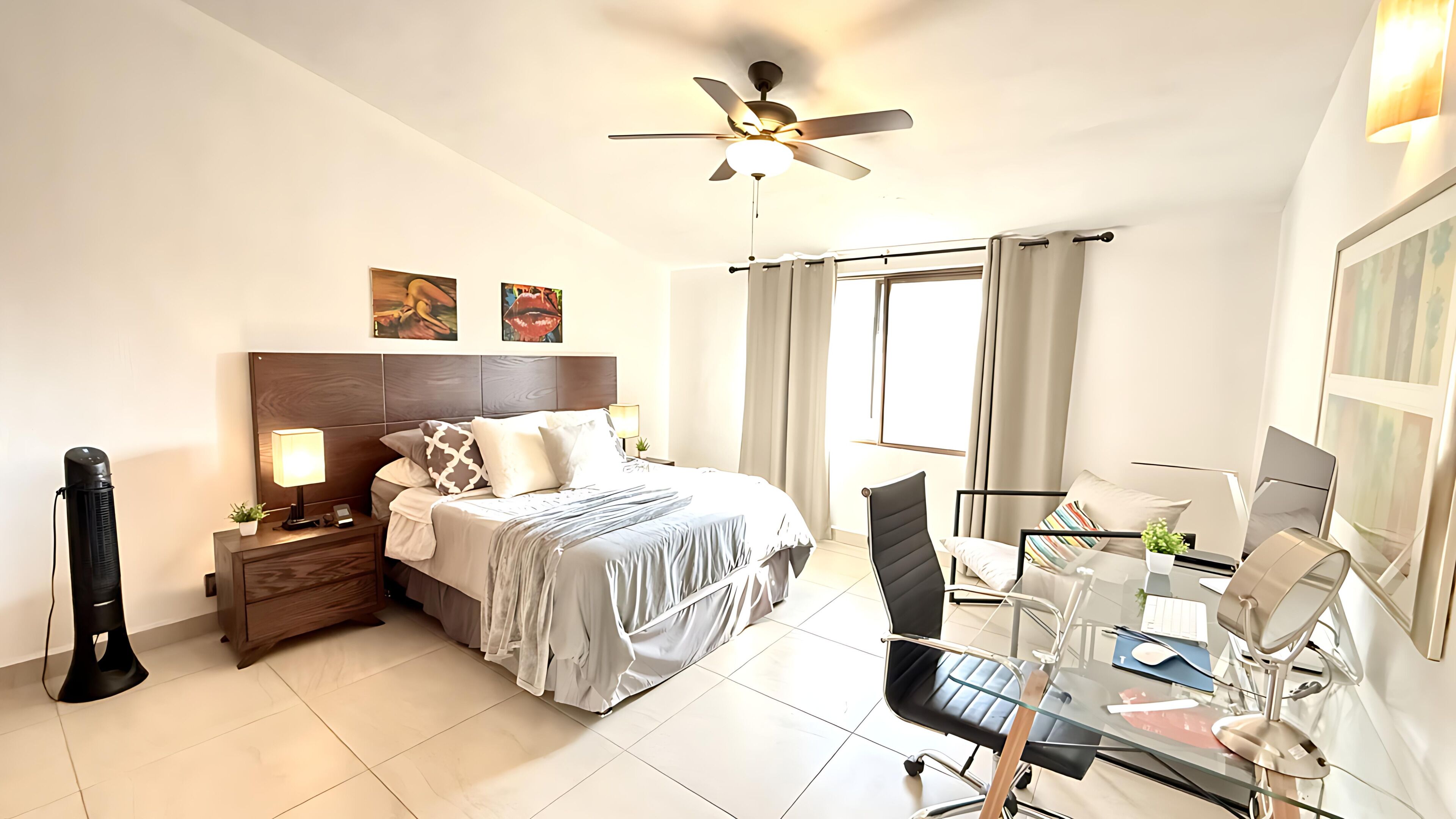 Habitación de hotel con cama, escritorio, silla, ventilador de techo y ventana con cortinas.