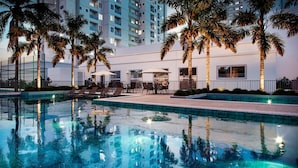 Piscina con sillones de descanso y sombrillas, rodeada de palmeras y un edificio con balcones.