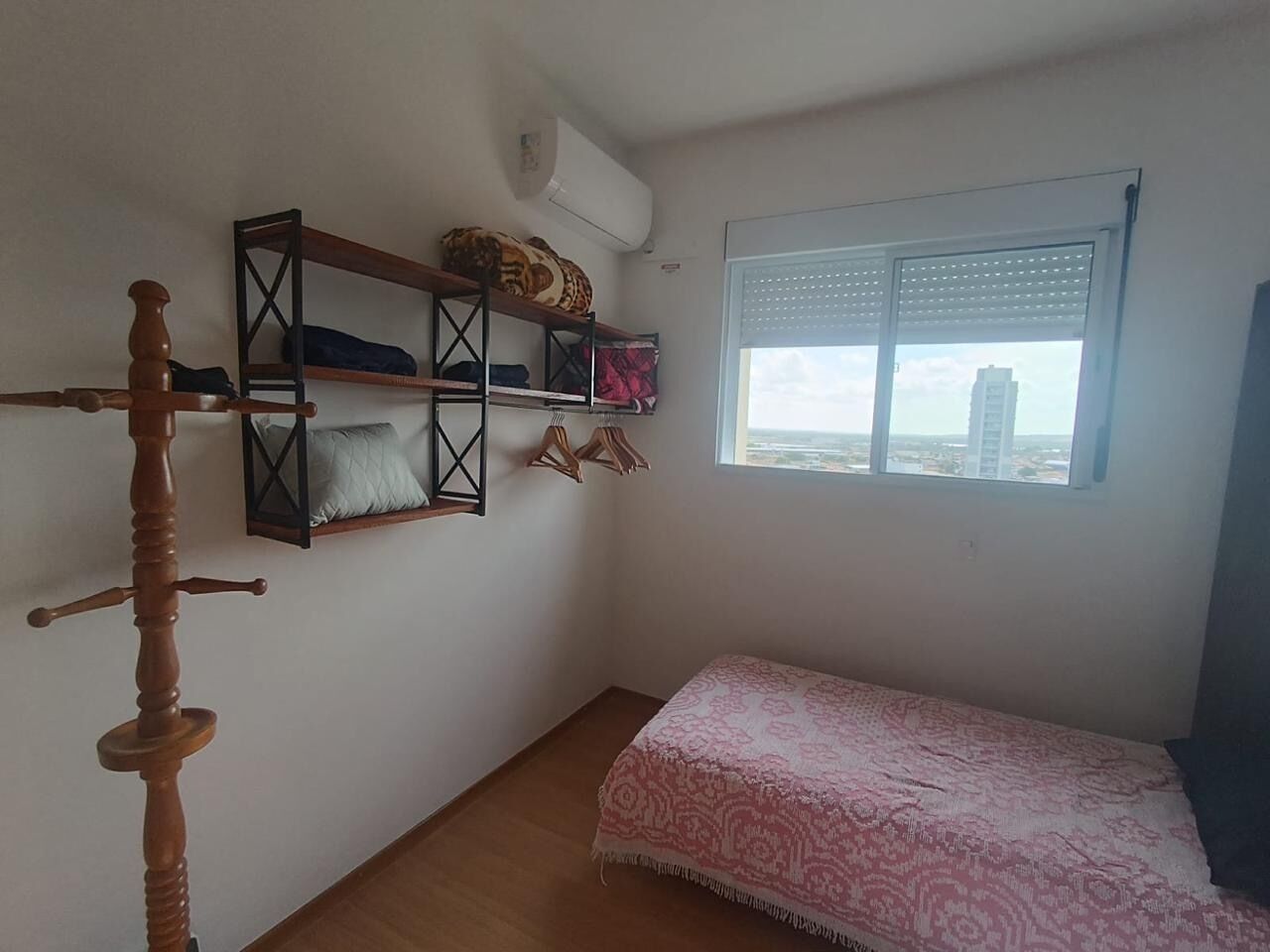 A small bedroom with a single bed, a wooden wardrobe, a window with blinds, and a view of buildings outside.