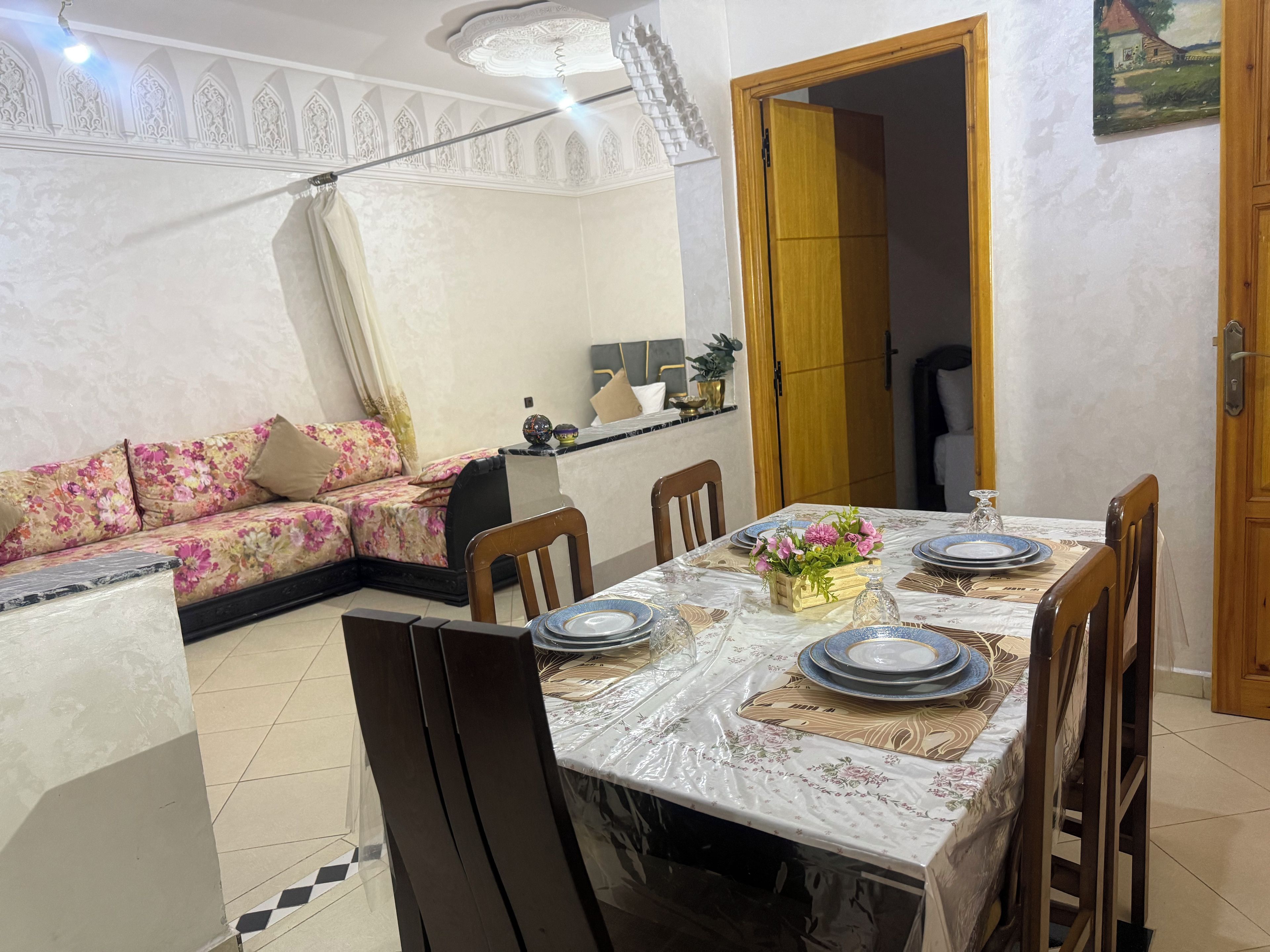 A dining area with a table set for four, surrounded by chairs, with a floral sofa in the background.