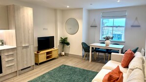 Un salon moderne avec un téléviseur à écran plat, une table à manger avec des chaises, un tapis vert et un canapé avec des coussins orange.