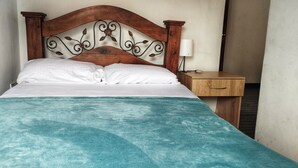 Una cama con cabecera de madera, ropa de cama blanca y una manta verde azulado. Hay una mesita de noche con una lámpara y un objeto pequeño encima.