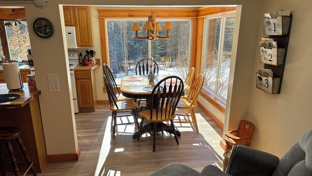 A dining area with a wooden table and chairs, a chandelier, and a view of trees through large windows. - True Adirondack Retreat! Nestled in the Mountains! (Mayfield)
