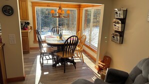 A dining area with a wooden table and chairs, a chandelier, and a view of a snowy landscape through large windows. - True Adirondack Retreat! Nestled in the Mountains! (Mayfield)
