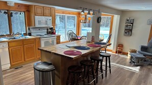 A kitchen with wooden cabinets, a stainless steel trash can, a bar area with stools, and a view of a snowy landscape through large windows. - True Adirondack Retreat! Nestled in the Mountains! (Mayfield)