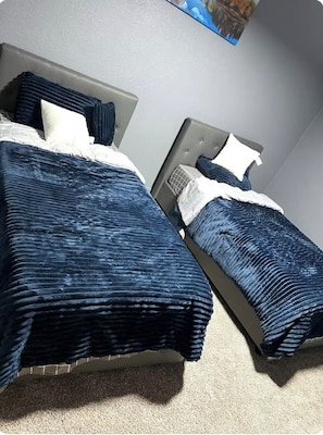 Two single beds with grey headboards and dark blue bedding are positioned against a light wall, accompanied by a painting.