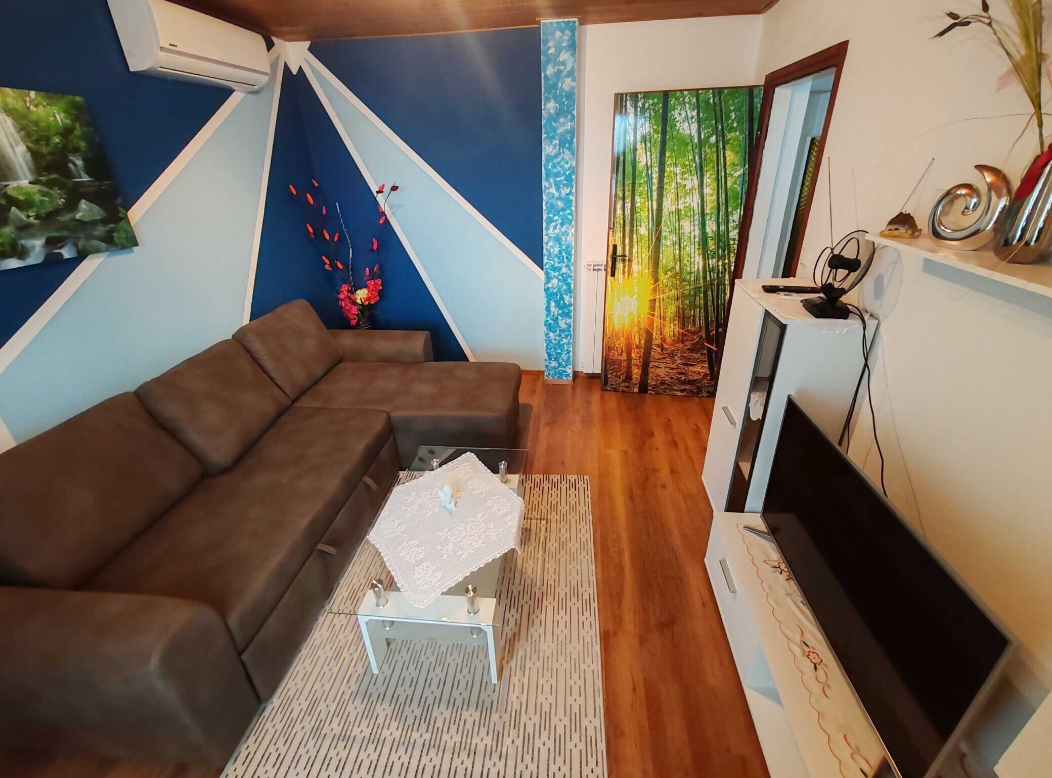 Un salon moderne avec un canapé d’angle, un téléviseur à écran plat et un mur décoratif représentant un paysage forestier.