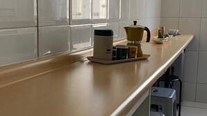 A kitchen with a long countertop, a coffee maker, a kettle, and a coffee pot. There are stools and a window with a view of buildings.