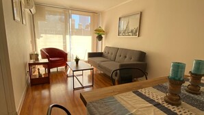 A living room with a sofa, a dining table with chairs, a red chair, a floor lamp, and a window with a cityscape view.