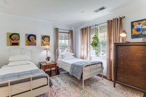 Une chambre avec deux lits, une commode, une fenêtre avec des rideaux et des œuvres d’art accrochées au mur.