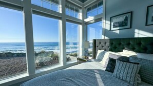 Un dormitorio con un ventanal grande con vistas al océano, una cama con ropa de cama estampada y una mesita de noche con una lámpara.