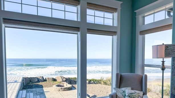 Una habitación con grandes ventanales que dan a una playa, un sillón con cojines y una lámpara de pie.