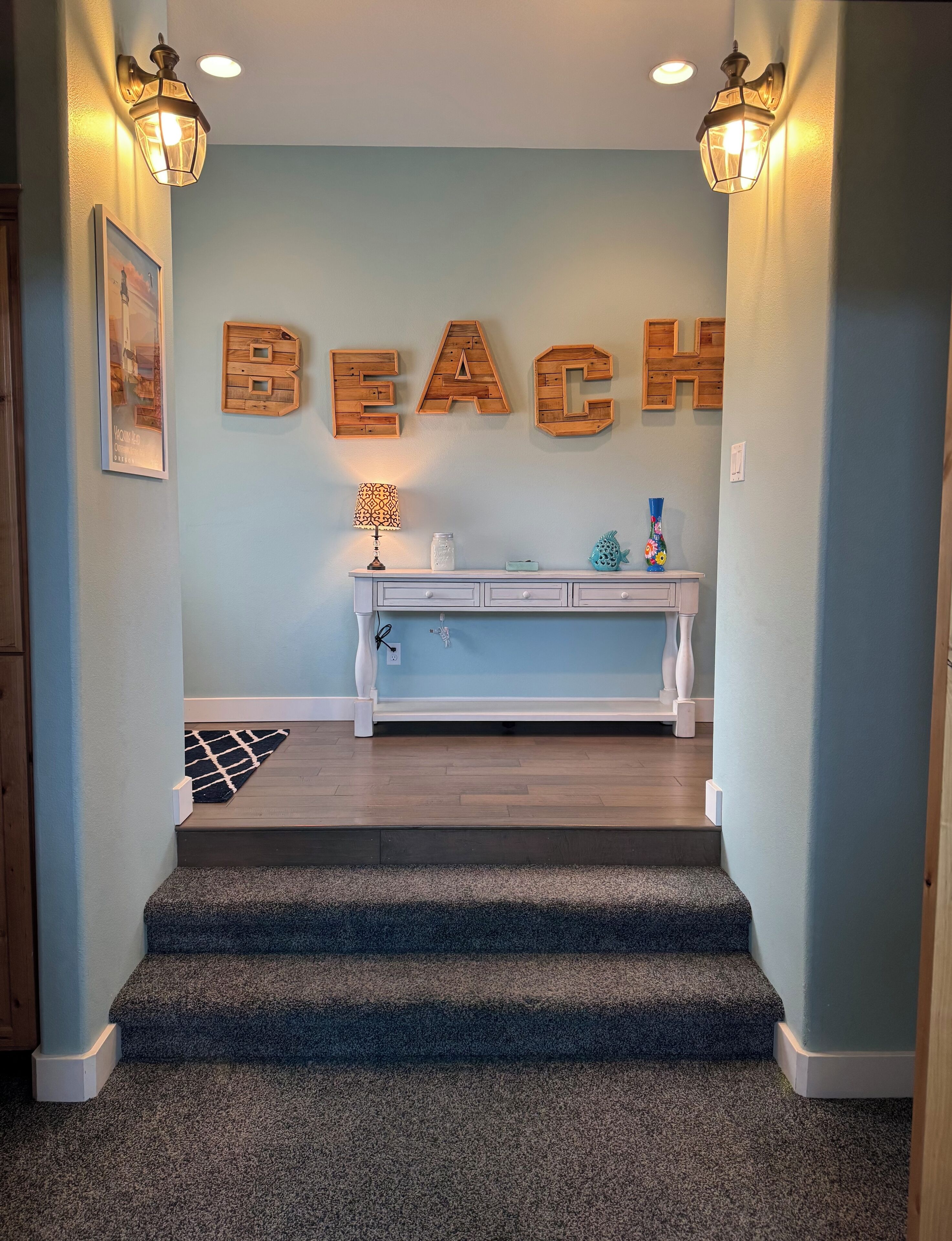 Un escalier menant à un palier avec un mur bleu clair, des lettres en bois formant le mot « PLAGE », une console blanche ornée d’objets décoratifs et un tapis noir et blanc.