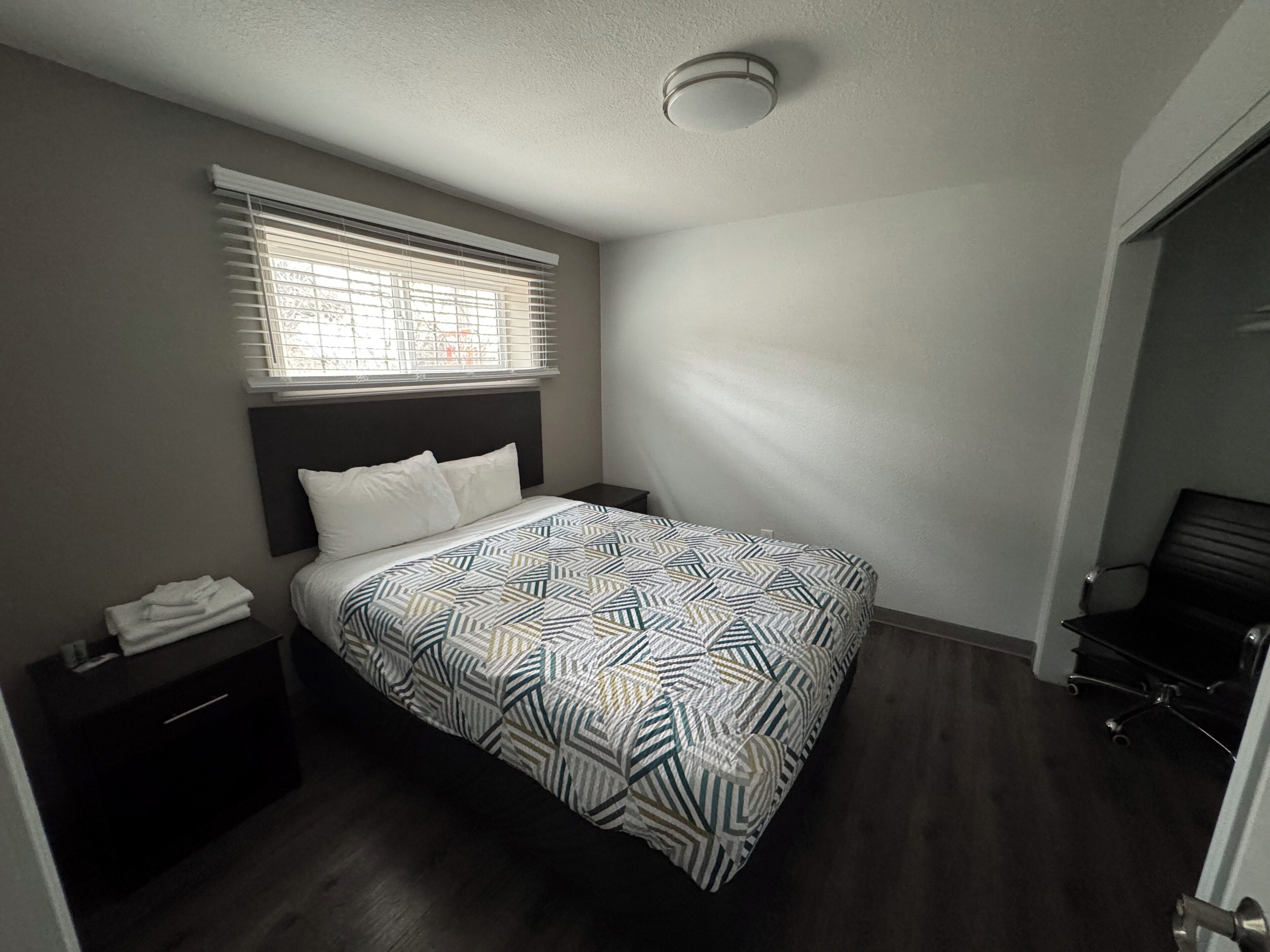 A bedroom with a bed, a nightstand, a window with blinds, a ceiling light, and a door.