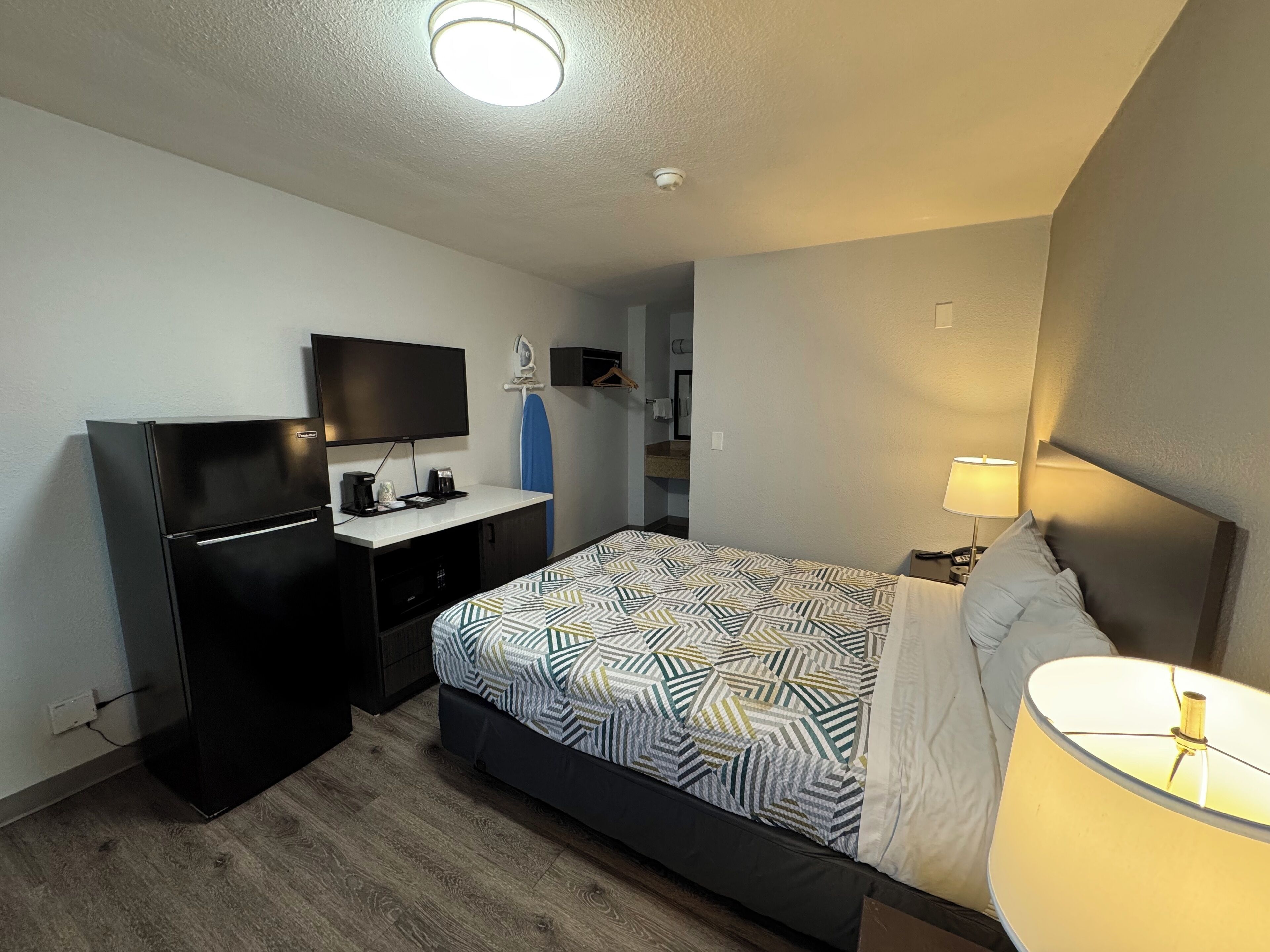 A hotel room with a bed, a desk with a flat-screen TV, a small kitchenette, and a bathroom visible through an open door.