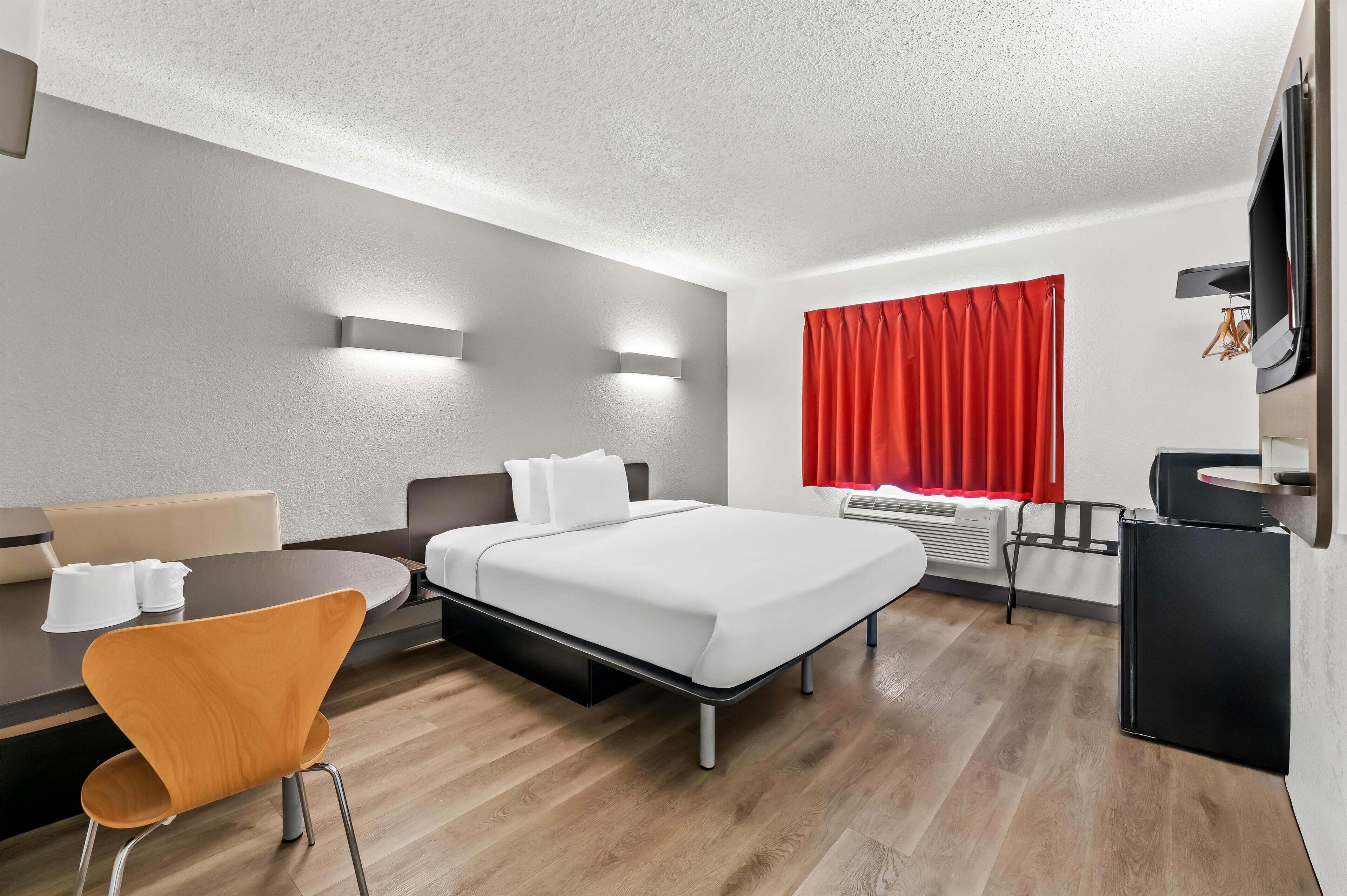 Une chambre d’hôtel moderne équipée d’un lit, d’un bureau, d’une chaise, d’une télévision et d’une fenêtre avec des rideaux rouges.