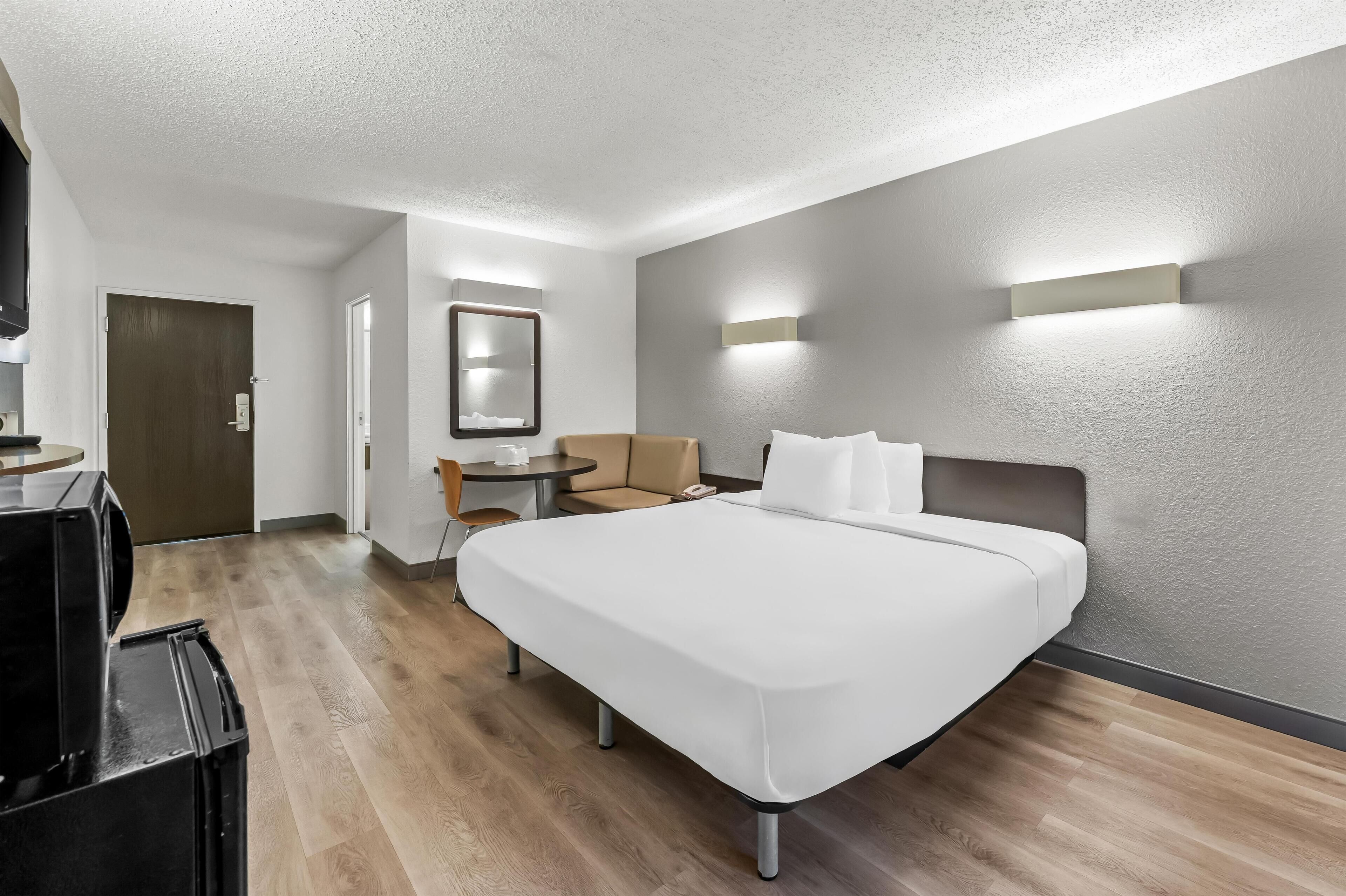 A hotel room with a large bed, a desk with a chair, a small table, a mirror, and a TV.