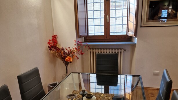 Een eetgedeelte met een glazen tafel, zwarte stoelen, een decoratief middelpunt en een raam met houten luiken.