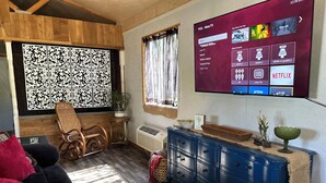 A living room with a wooden ceiling, a blue cabinet, a black sofa, and a large TV screen displaying a streaming service interface. - Charming 2-bedroom cottage in Dexter with AC, WiFi. Enjoy your stay (Dexter)