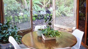 Een houten interieur met een ronde tafel en witte stoelen, omringd door glazen deuren met uitzicht op een weelderige tuin.