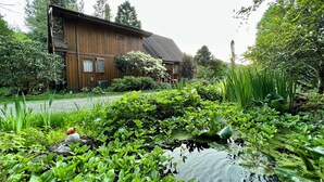 Een houten huis met een vijver op de voorgrond, omringd door weelderige begroeiing en bomen.