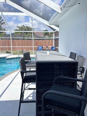 An outdoor patio with a pool, black wicker furniture, and a wooden fence in the background. - Charming Coastal Vibe, 3BR Home with Heated Pool (Cape Coral)