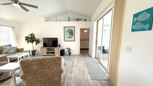 A spacious living room with a television, sofa, and coffee table. There is a ceiling fan, a potted plant, and a sliding glass door leading to a balcony. - Charming Coastal Vibe, 3BR Home with Heated Pool (Cape Coral)