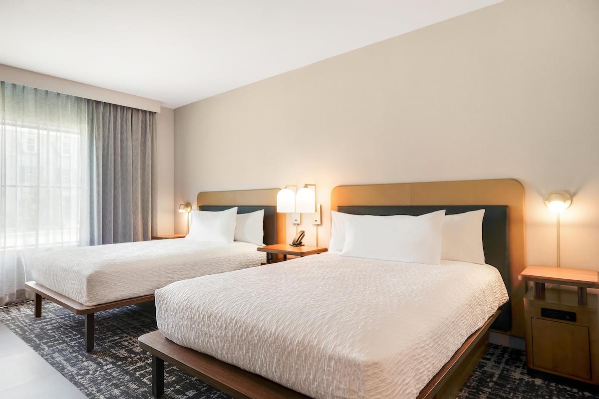 A hotel room with two beds, a bedside table with a lamp, and a large window with curtains.