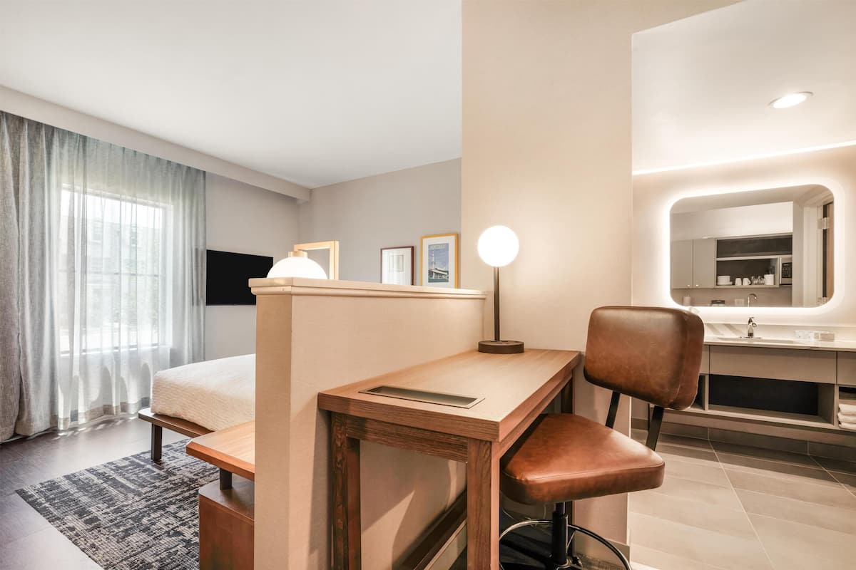 A modern hotel room with a bed, desk, chair, and a bathroom with a large mirror.