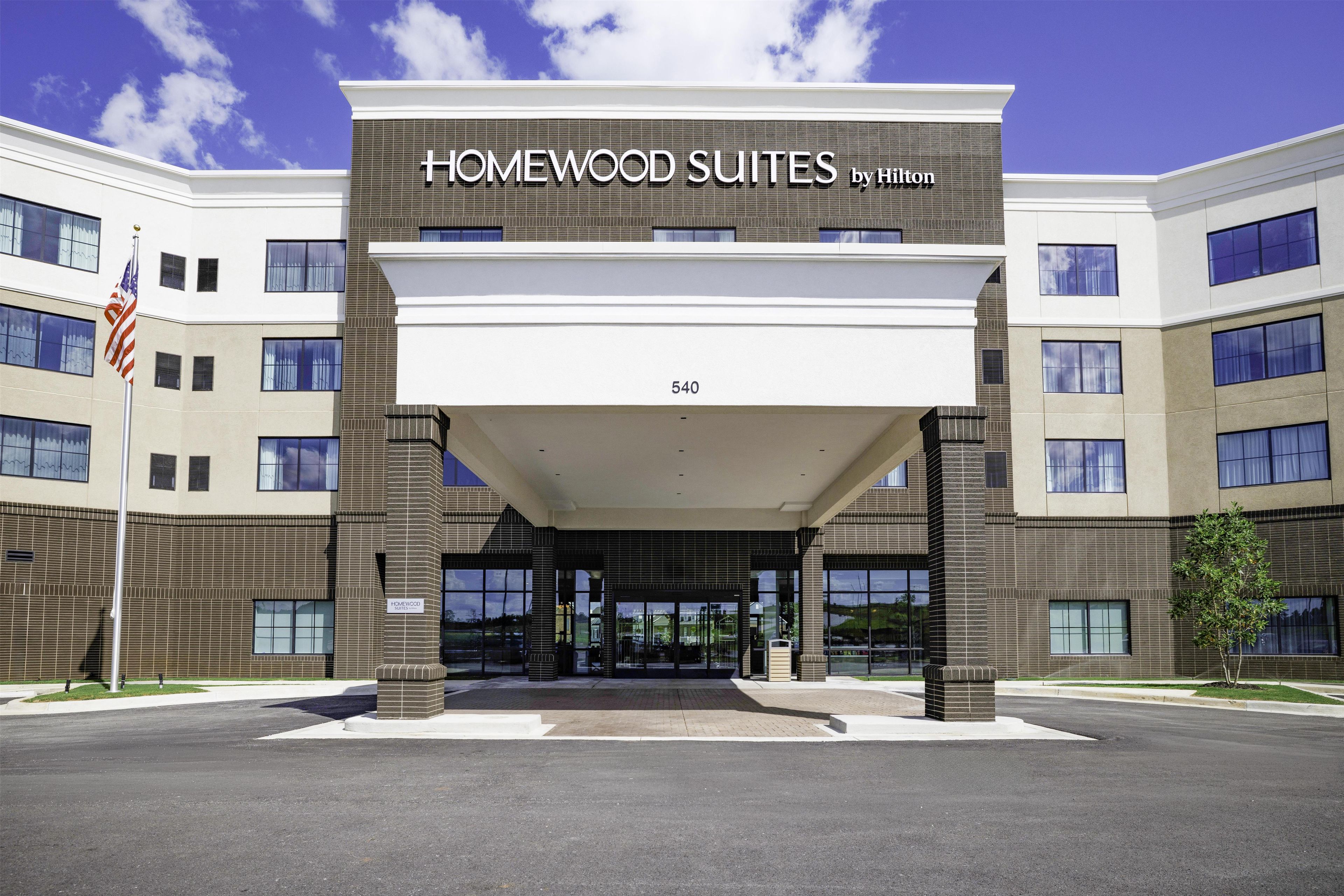 A hotel entrance with 'Homewood Suites by Hilton' signage, a flag, and a clear sky.