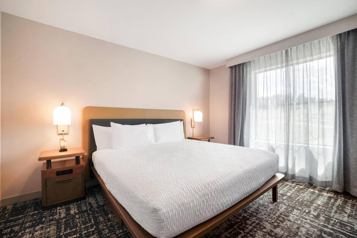 A hotel room with a large bed, two bedside tables with lamps, a bench at the foot of the bed, and a large window with curtains.