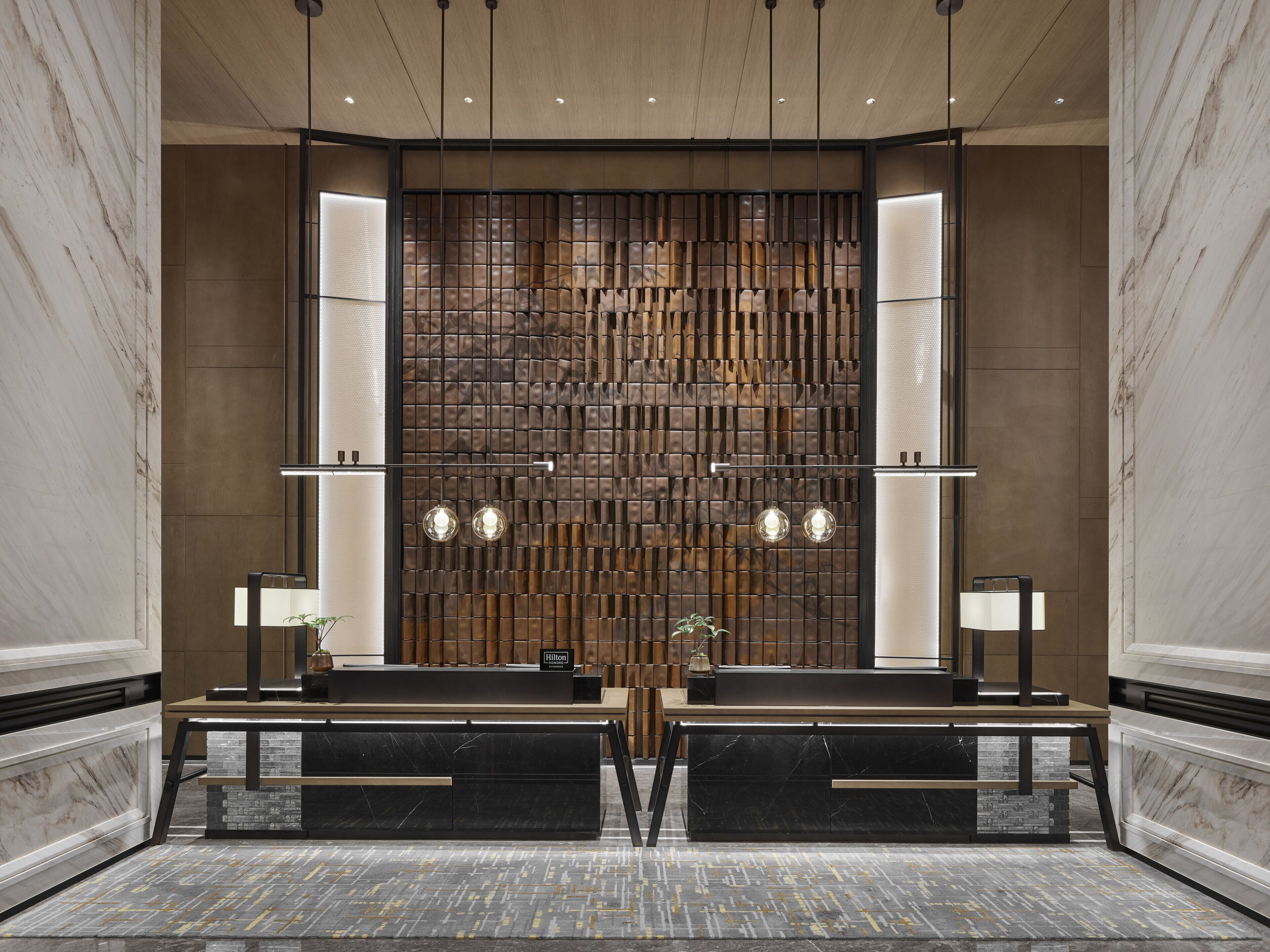 A hotel reception area with marble walls, a wooden panel wall, two reception desks, and hanging pendant lights.