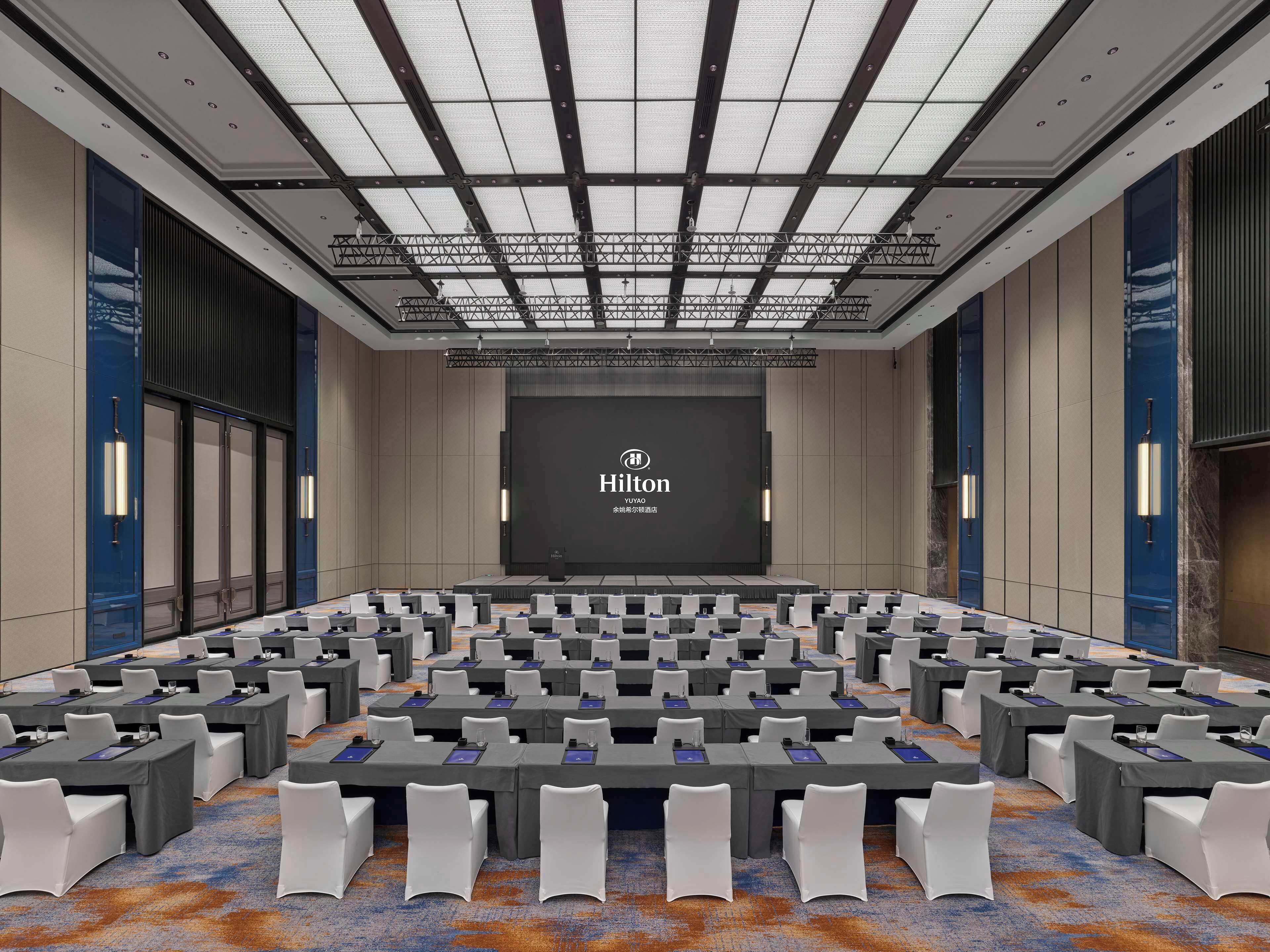 Kokoushuone, jossa on suuri näyttö, jossa näkyy Hiltonin logo, useita pöytiä ja tuoleja riveissä sekä kuvioitu matto.