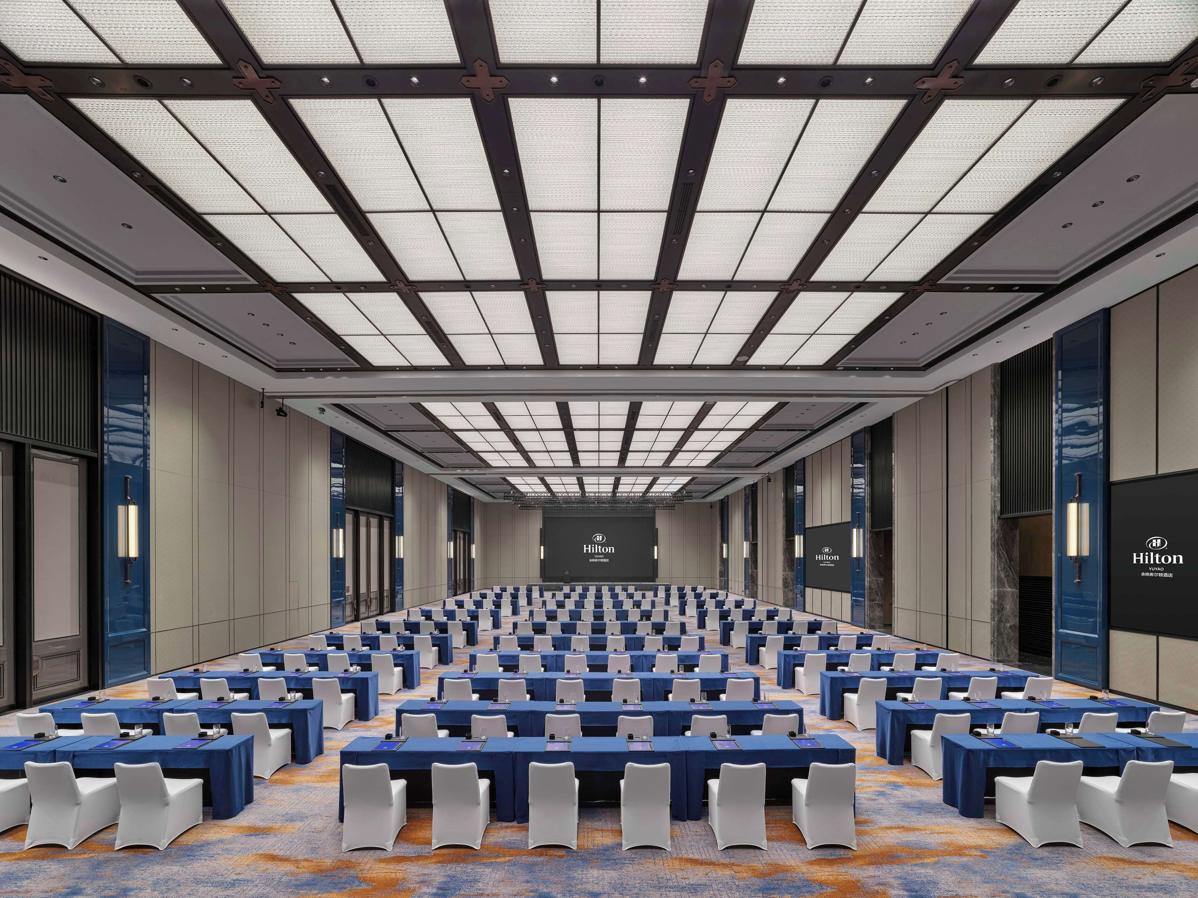 A large conference room with rows of tables and chairs, a high ceiling with a grid pattern, and a large screen at the front.