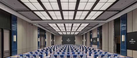 A large conference room with rows of tables and chairs, a high ceiling with a grid pattern, and a large screen at the front.