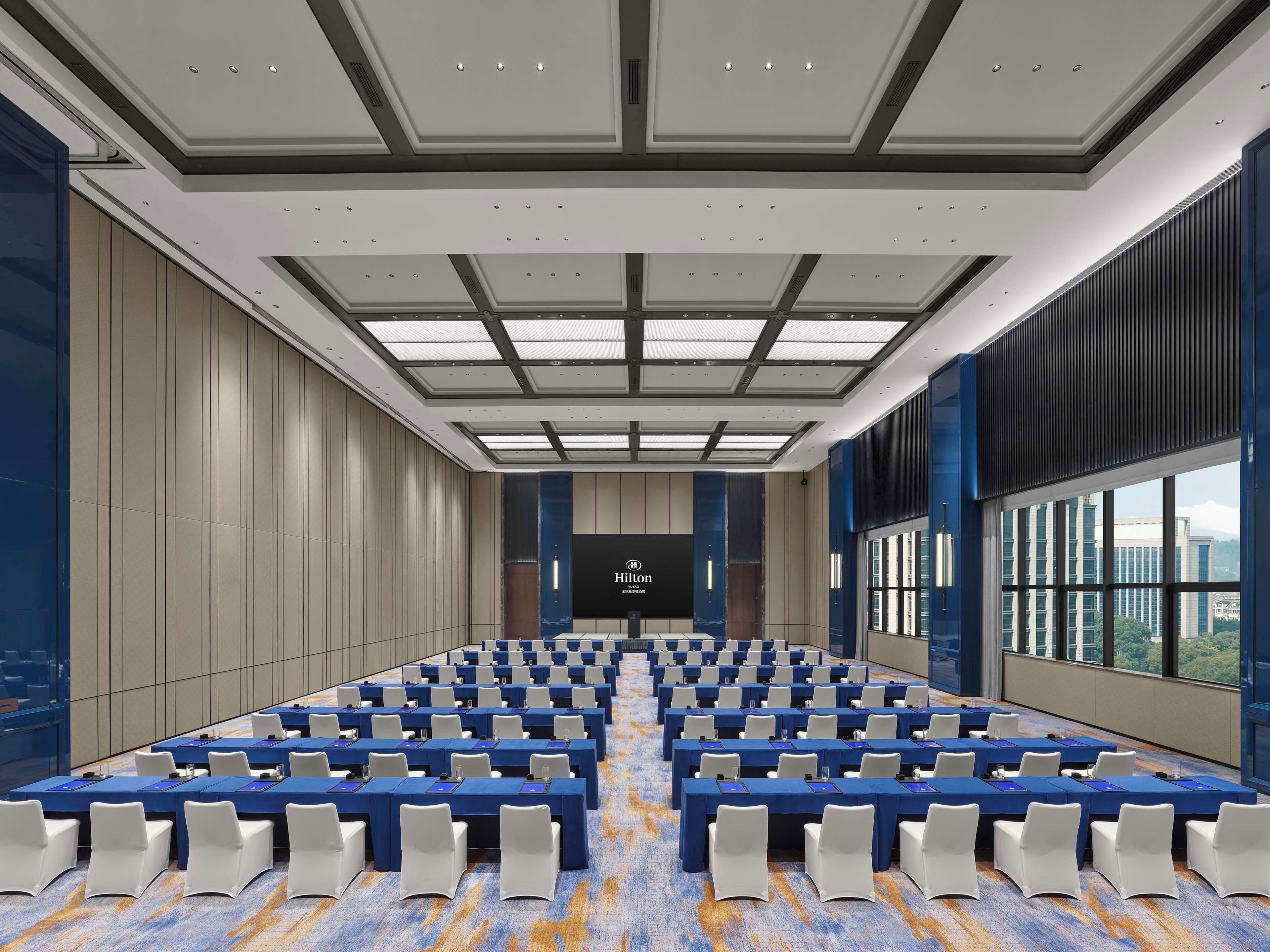 A large conference room with rows of tables and chairs, a high ceiling with recessed lighting, and a view of buildings through large windows.