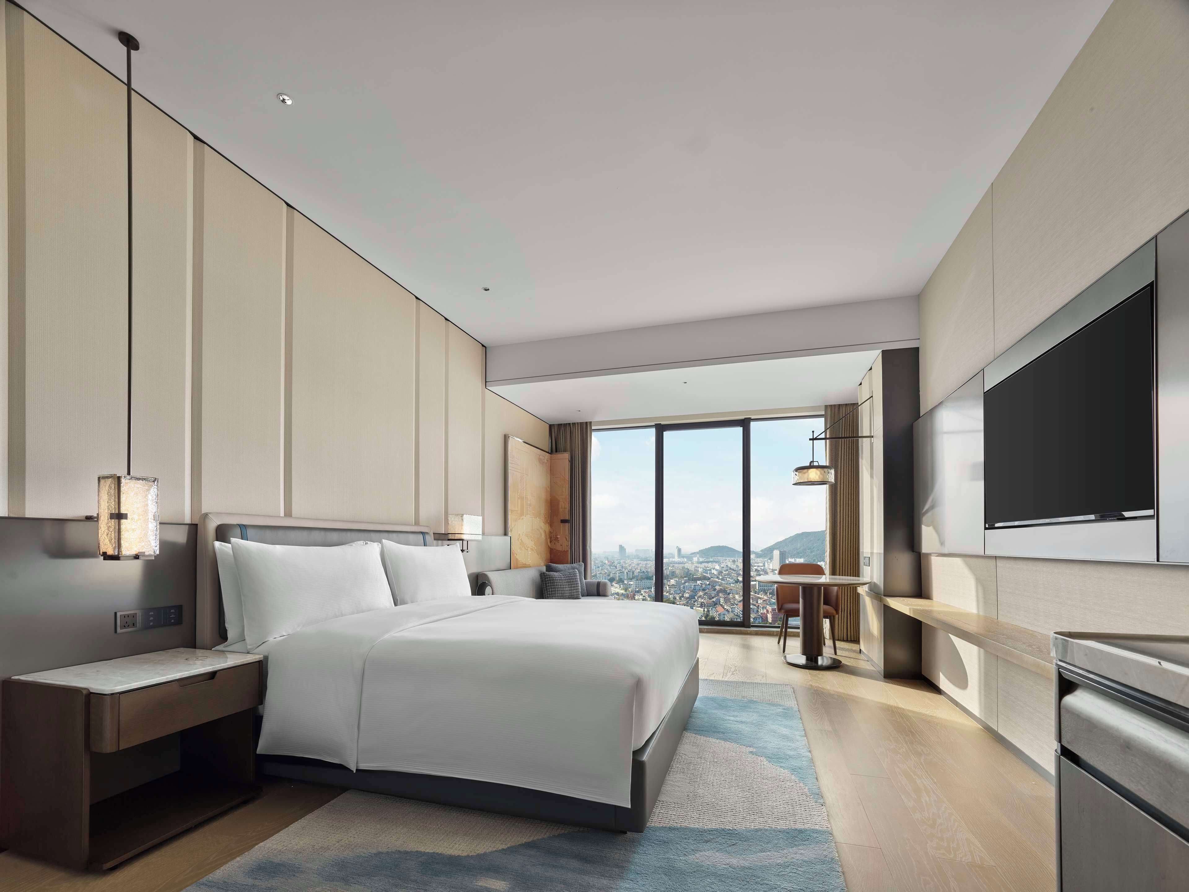 Une chambre d’hôtel moderne dotée d’un grand lit, de tables de chevet, d’un bureau, d’une télévision et offrant une vue sur la ville grâce à de grandes fenêtres.