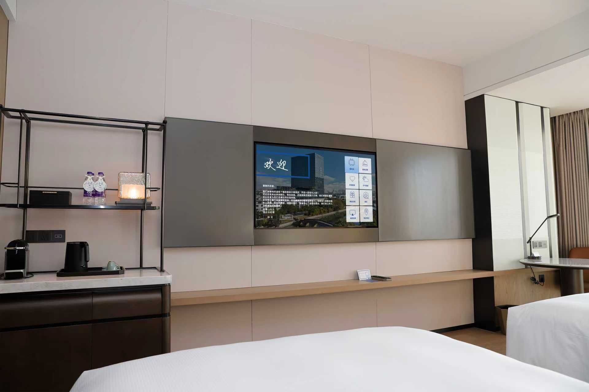 A hotel room with a large flat-screen TV mounted on the wall, a desk with a coffee maker, and a bed with white bedding.