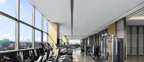 A modern gym with cardio machines, weight stations, and a large window offering a city view.