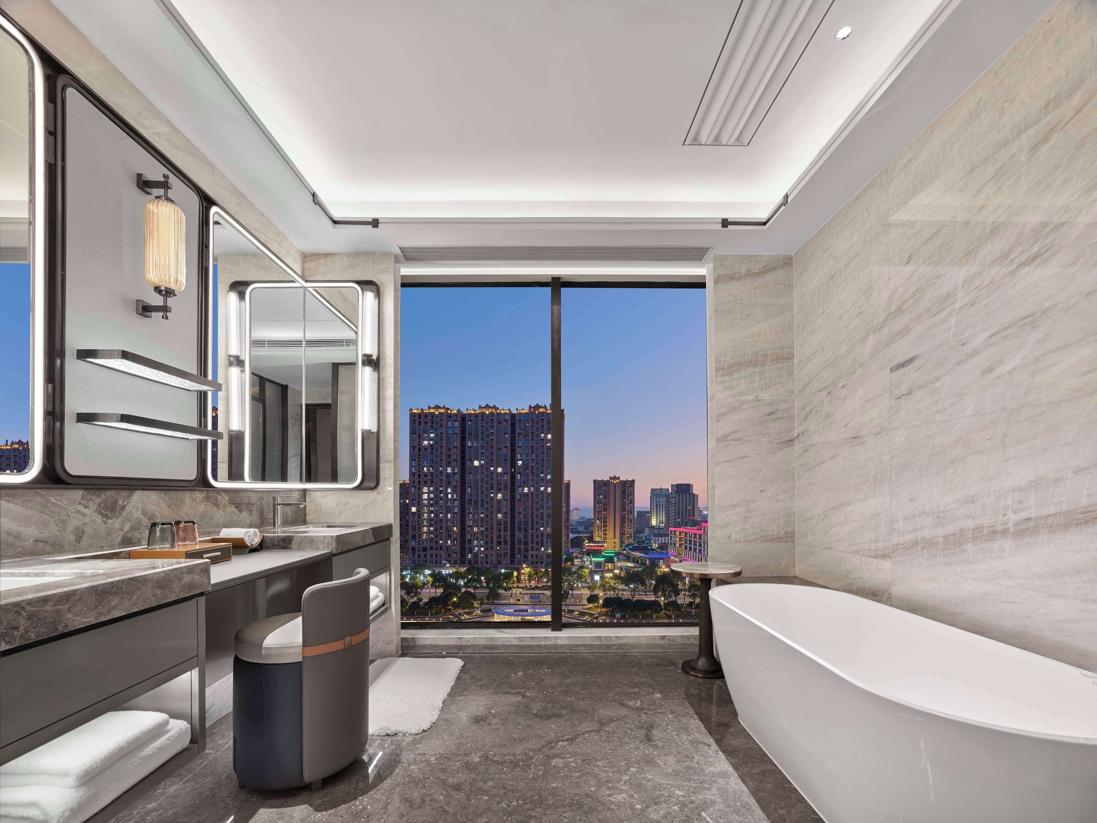 A modern bathroom with a large mirror, a bathtub, and a view of a cityscape at dusk.