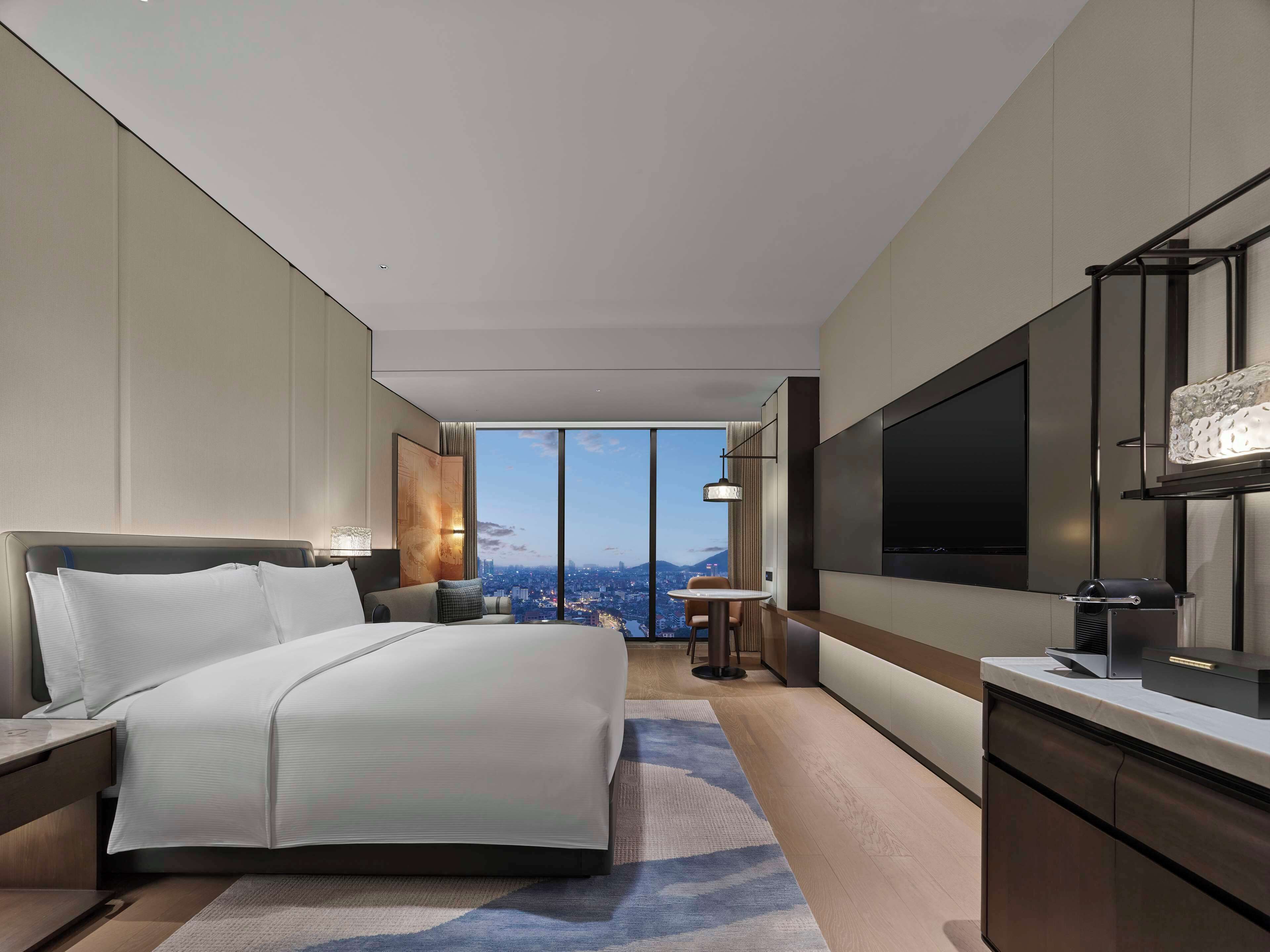 A modern hotel room with a large bed, a TV, a desk, and a view of the city.