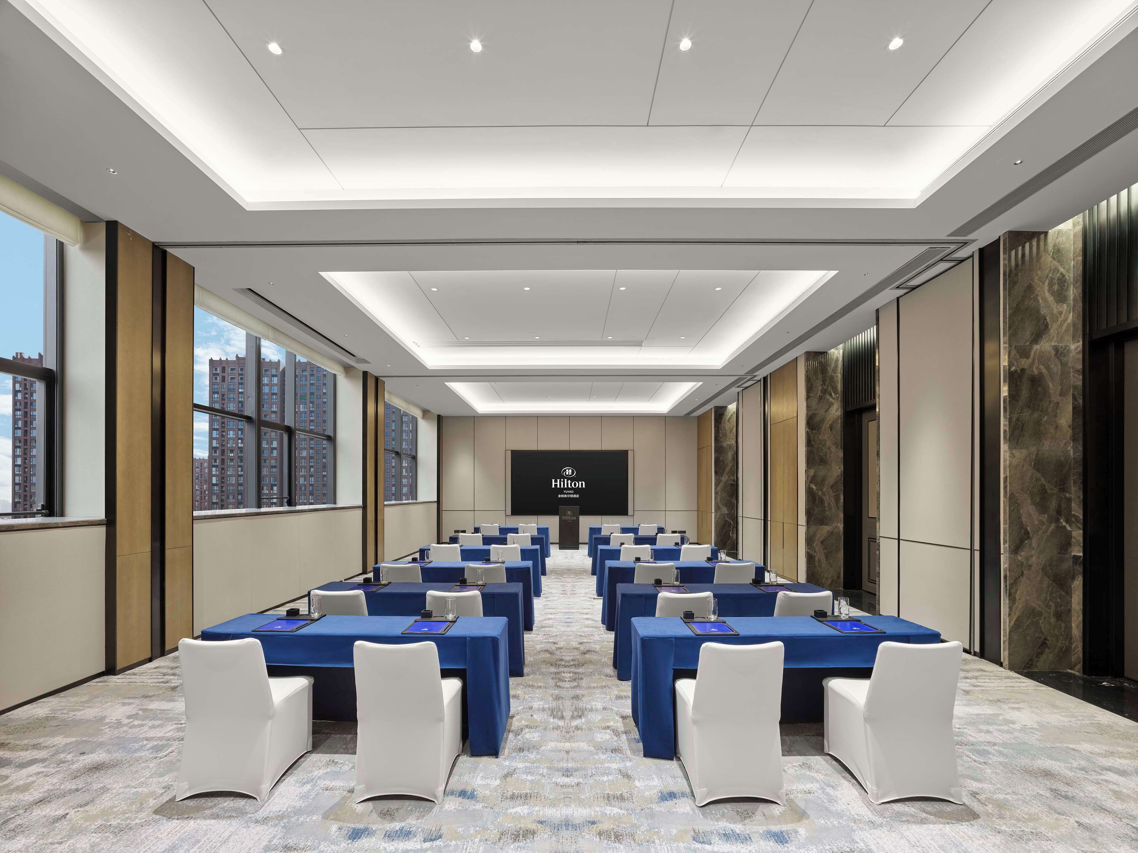 A conference room with multiple tables and chairs, a large screen at the front, and large windows with a city view.