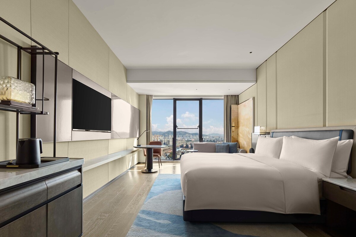 A modern hotel room with a large bed, a flat-screen TV mounted on the wall, a desk with a coffee maker, and a city view through large windows.