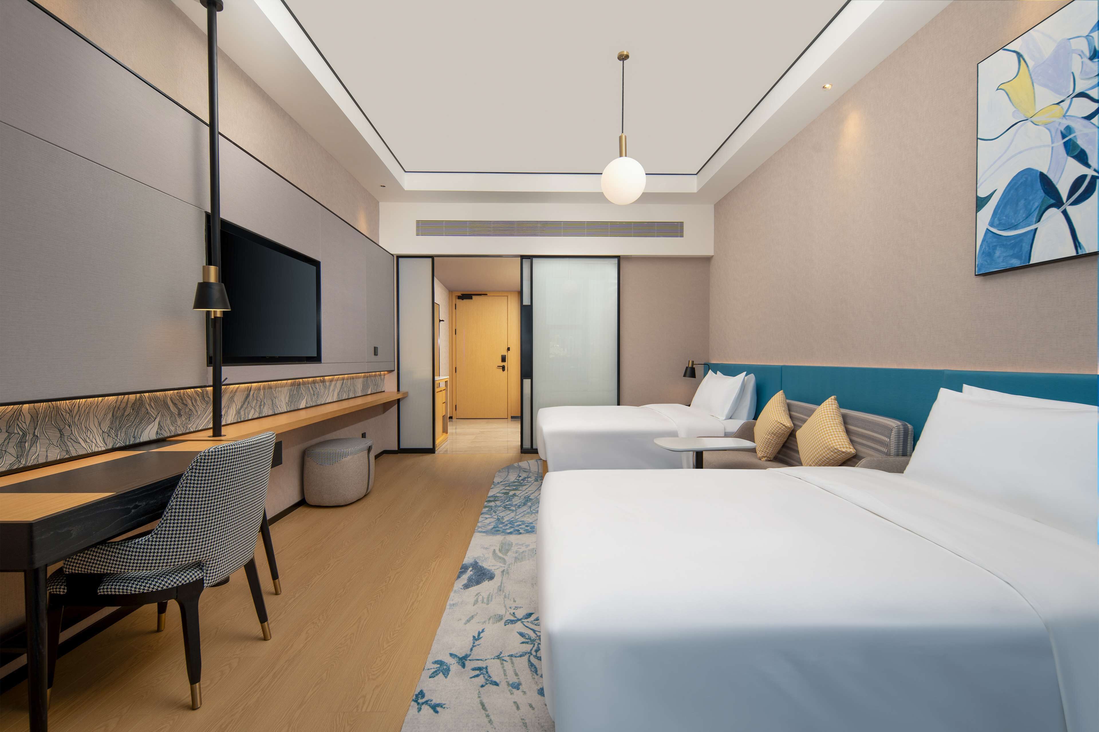 Habitación de hotel moderna con televisor de pantalla plana, un escritorio con silla, dos camas con ropa blanca y un cuadro abstracto grande en la pared.