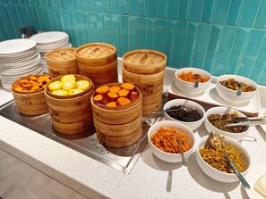 Buffet com diversos alimentos, incluindo pãezinhos cozidos no vapor, frutas fatiadas e legumes variados.