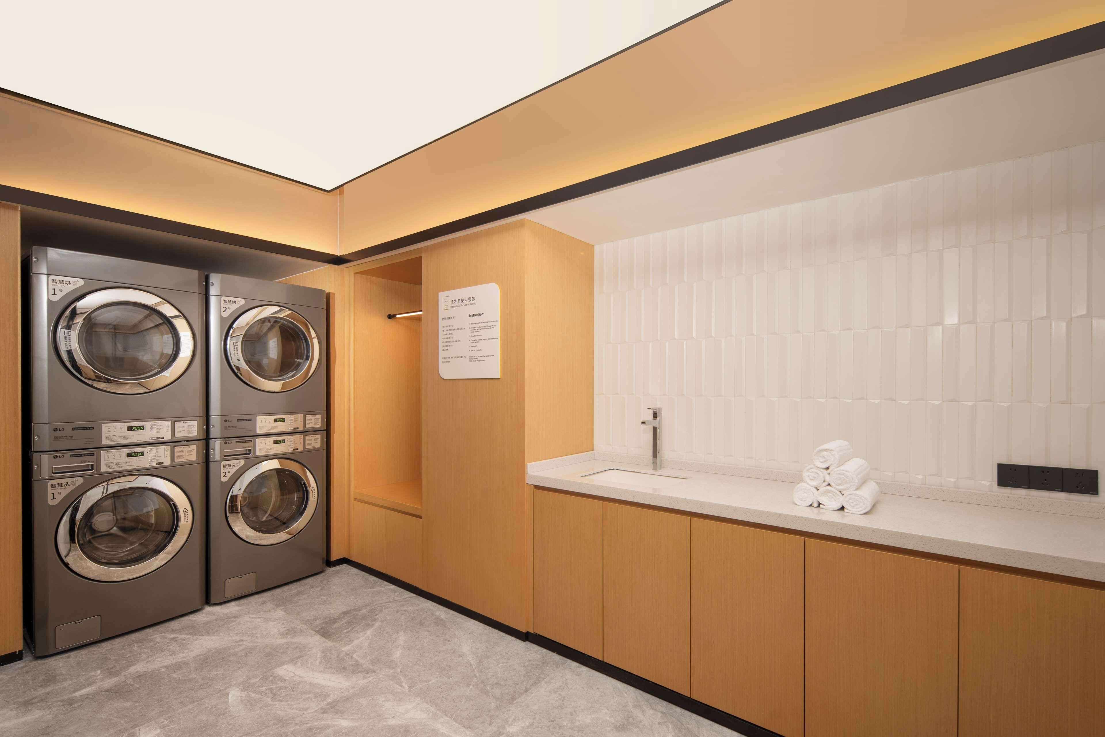 A modern laundry room with two stacked front-loading washing machines, a sink, and a white tiled wall.