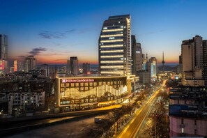 A modern hotel with a prominent glass facade and illuminated signage, surrounded by a cityscape at dusk. - Hilton Garden Inn Xuzhou Pengcheng Square (Xuzhou)