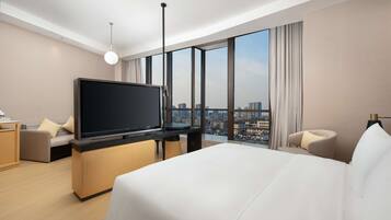 A modern hotel room with a large bed, a flat-screen TV, a desk, and a view of the cityscape through large windows.